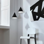 Two black cone pendants from Clérachic hang beside a tall white wall with a bold black-and-white painting and a white chair.
