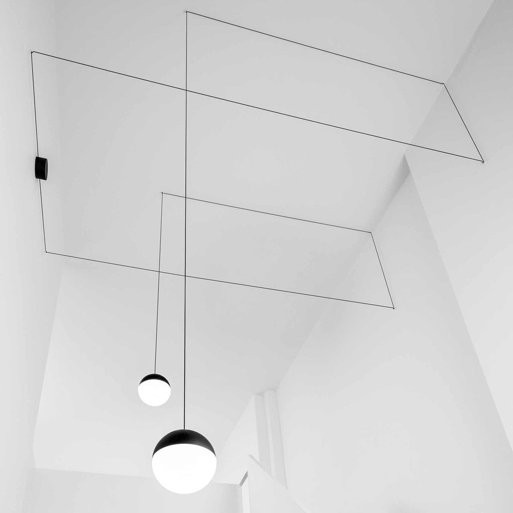Looking up at a Clérachic pendant with a black and opal sphere shade, its thin cable running in angled lines across a white ceiling.