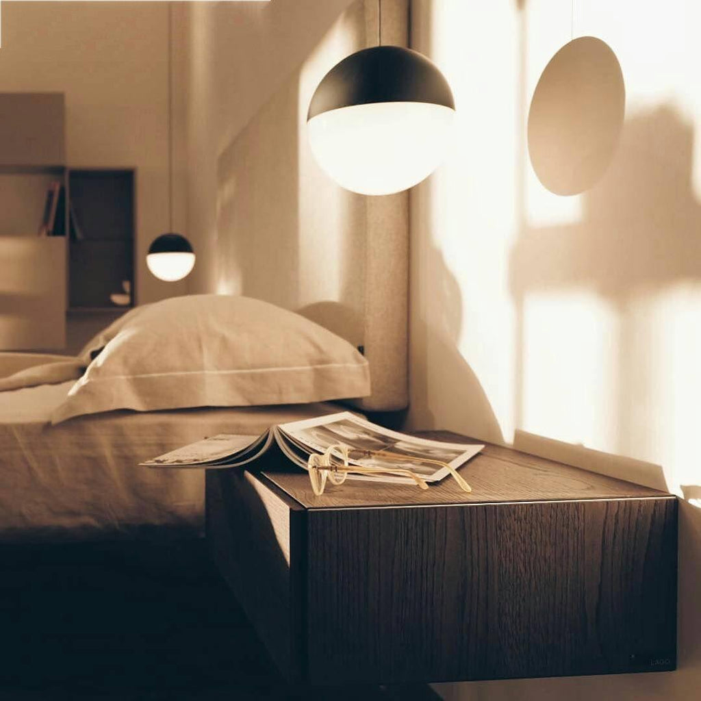Warm evening light falls across a bedside, with a Clérachic pendant with a half black, half opal sphere shade above the bed.