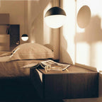 Warm evening light falls across a bedside, with a Clérachic pendant with a half black, half opal sphere shade above the bed.