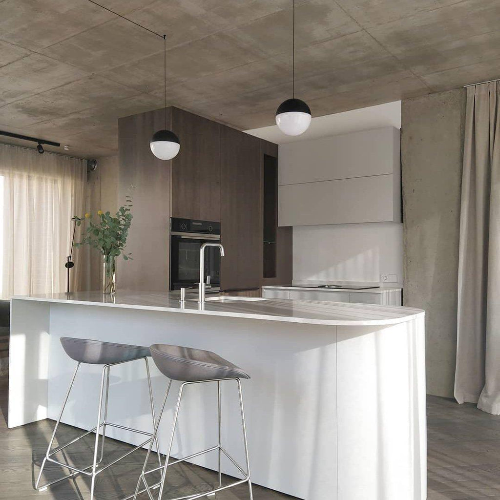 Two Clérachic pendants with half black, half opal sphere shades hang over a white kitchen island with grey stools and a concrete ceiling.