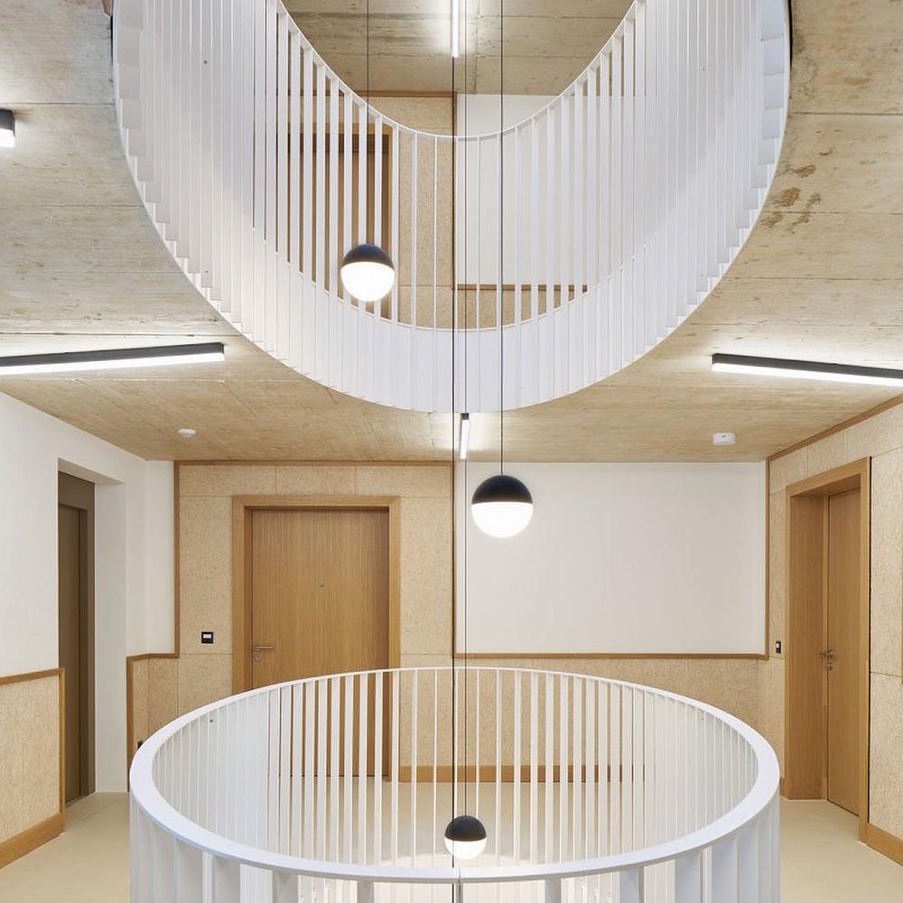 Clérachic sphere pendants hang down a stairwell with curved white balustrades and pale timber walls, seen from above.