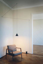 A single Clérachic cone pendant in black hung low over an armchair, its cable traced along the cream wall and ceiling edge.
