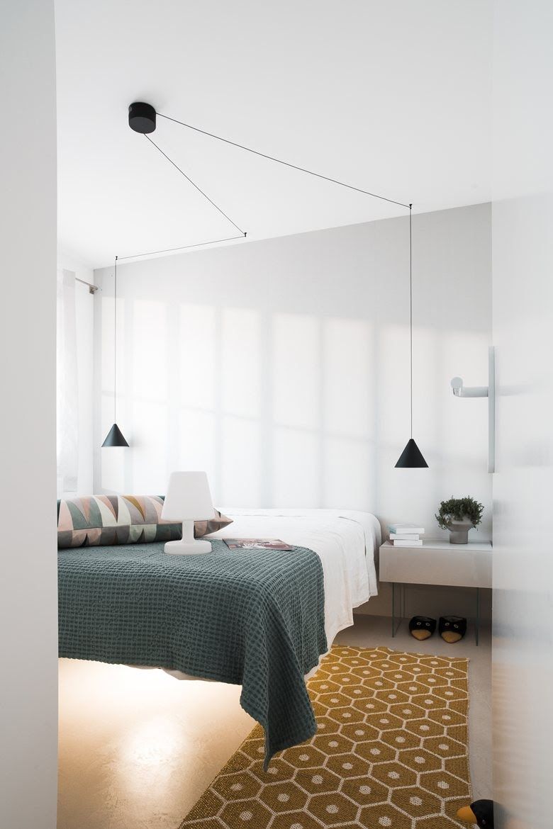 Two Clérachic black cone pendants hang at the foot of a bed with a green blanket, their cables running along a white wall.