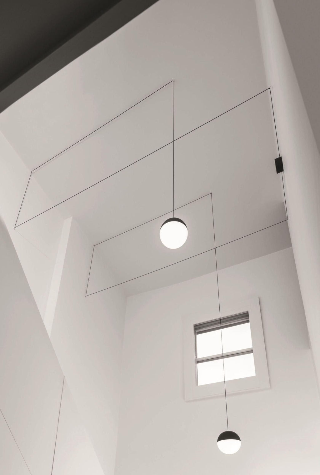 Looking up a tall white stairwell at two Clérachic sphere pendants on fine cables, with a small window below.