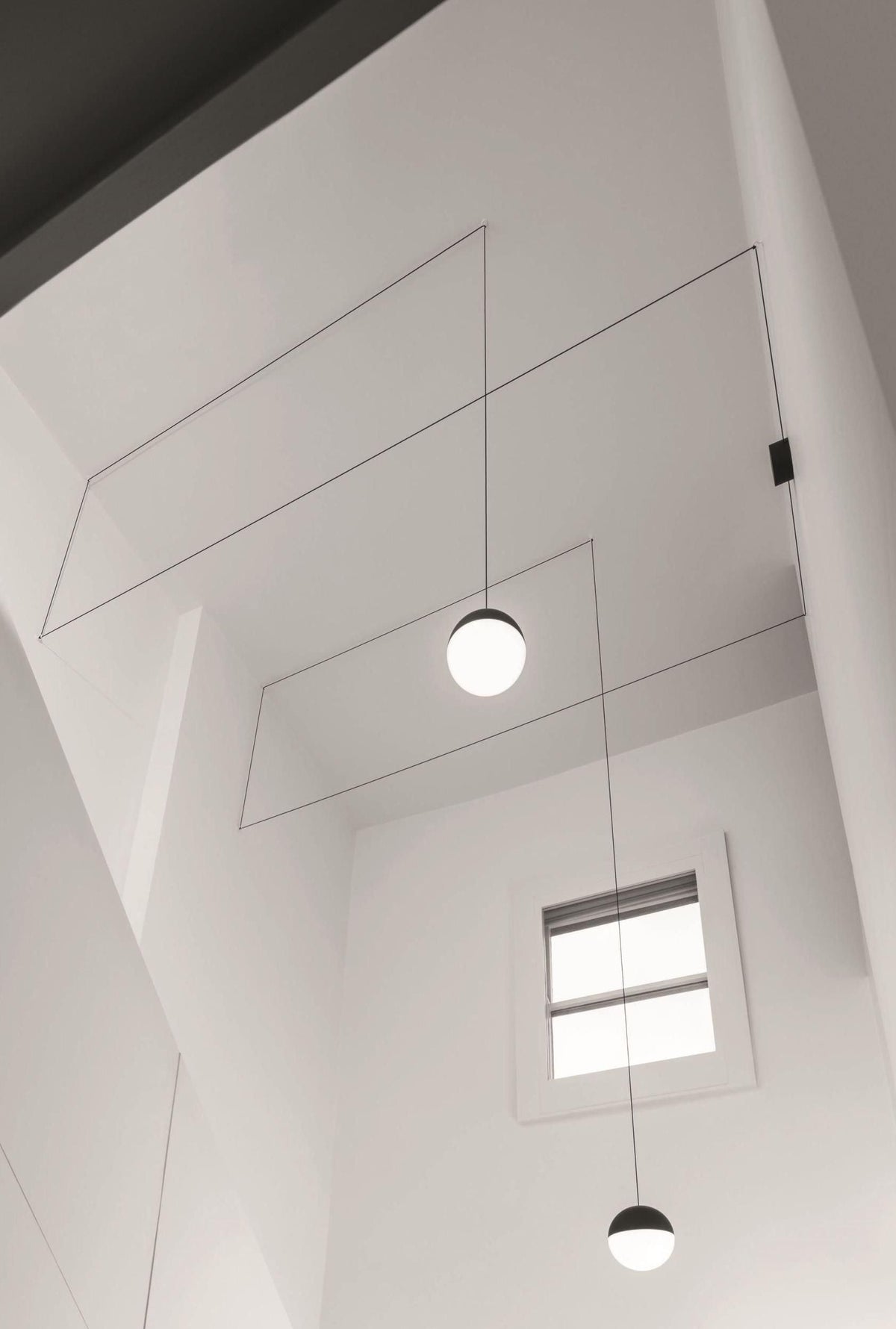Looking up a tall white stairwell at two Clérachic sphere pendants on fine cables, with a small window below.