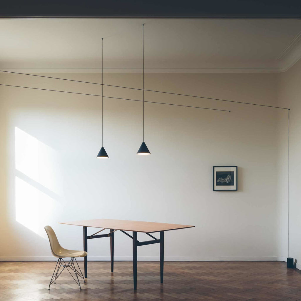 Two Clérachic black cone pendants on long drops beside a pale wall, with a wooden table, a moulded chair and herringbone floor.