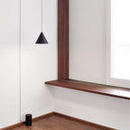 A Clérachic black cone pendant in a white room with a dark wood window frame, its cable running down the wall to a floor-level base.
