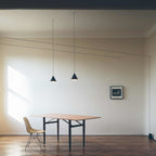 Same pair of Clérachic cone pendants in black, hung low in a bright room above a wooden table and a pale moulded chair.