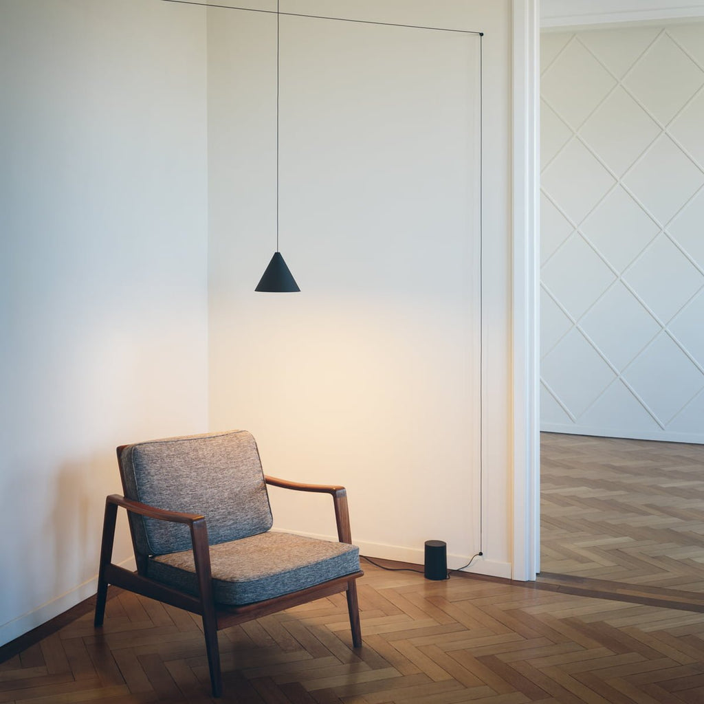 Clérachic cone pendant lit above a grey armchair with wooden arms, with its cable running down to a small black base on the floor.