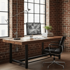 A Clérachic black adjustable lamp on a plank desk beside a monitor and a leather chair, in front of a brick wall and steel windows.