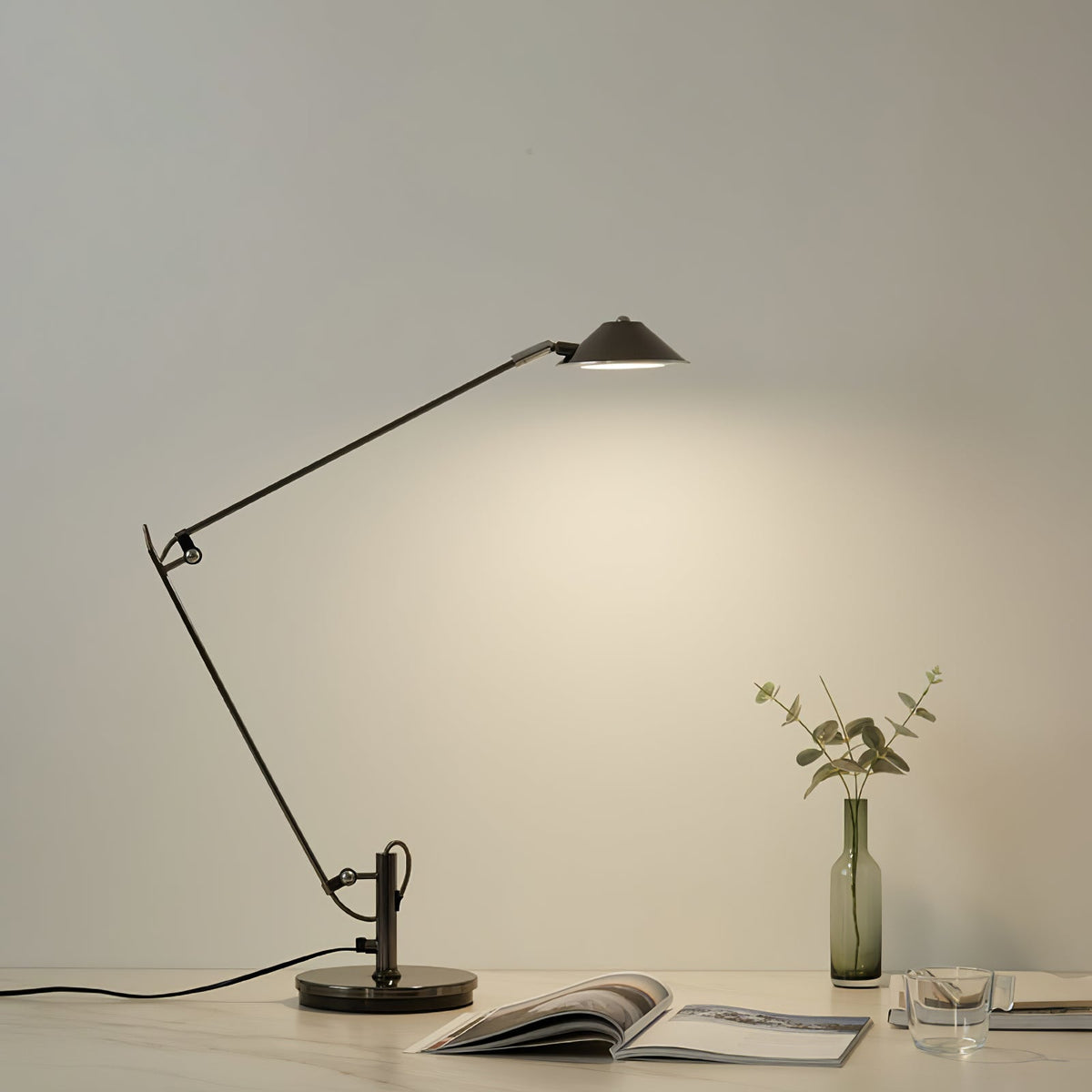 A Clérachic black arm lamp lights a cream wall from a pale table, next to a glass bottle of green stems and an open magazine.
