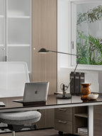 A Clérachic black adjustable lamp on a glossy desk beside a laptop and a wooden vase, with a window and plants beyond.