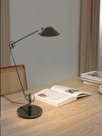 A Clérachic black arm lamp with a round base stands on a light oak desk, lighting an open book, with a rattan chair behind.