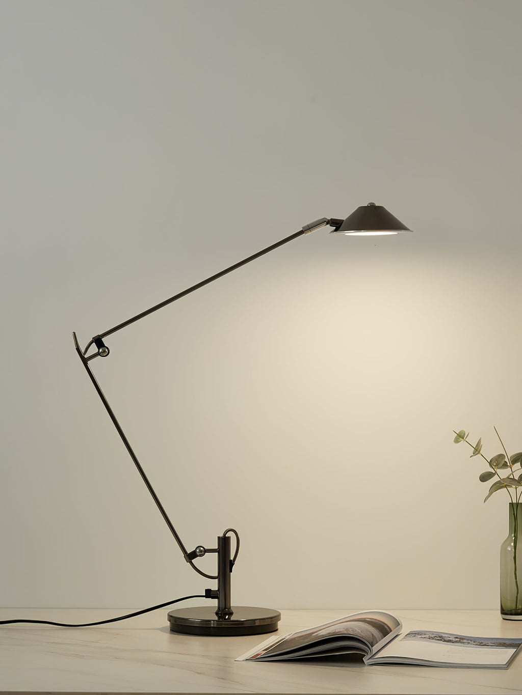 A Clérachic black arm lamp casts warm light on a pale wall from a desk, with an open magazine and a small bottle vase.