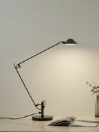 A Clérachic black arm lamp casts warm light on a pale wall from a desk, with an open magazine and a small bottle vase.