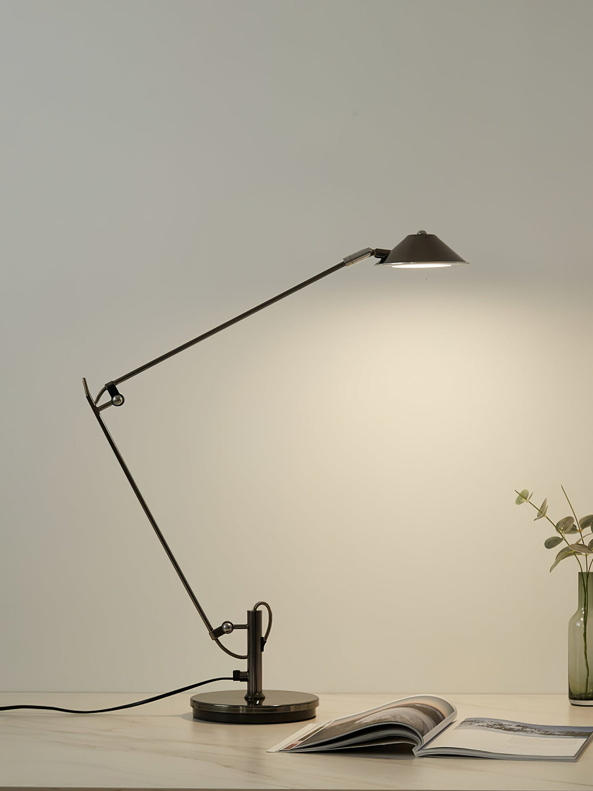 A Clérachic black arm lamp casts warm light on a pale wall from a desk, with an open magazine and a small bottle vase.