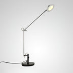 A Clérachic arm lamp in gunmetal black with its shade tilted up and lit, on a soft white background with the cord trailing left.