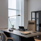A Clérachic black arm lamp at the far end of a dark office desk, in a room with a tall window and a leather chair.