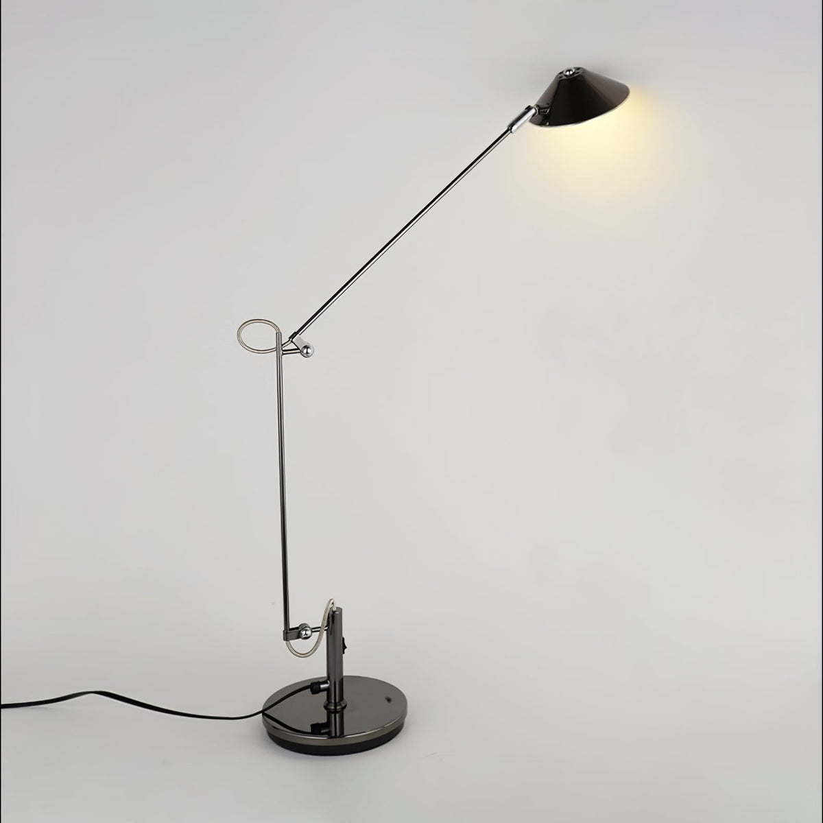 A Clérachic black arm lamp raised high with its shade angled down, casting a warm glow on a plain pale backdrop.