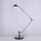 A Clérachic black arm lamp folded into a Z shape with its lit shade high on the left, on a soft lilac-grey backdrop.