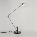 A Clérachic black adjustable lamp with its jointed arm folded and the lit shade tilted up on the right, on grey.