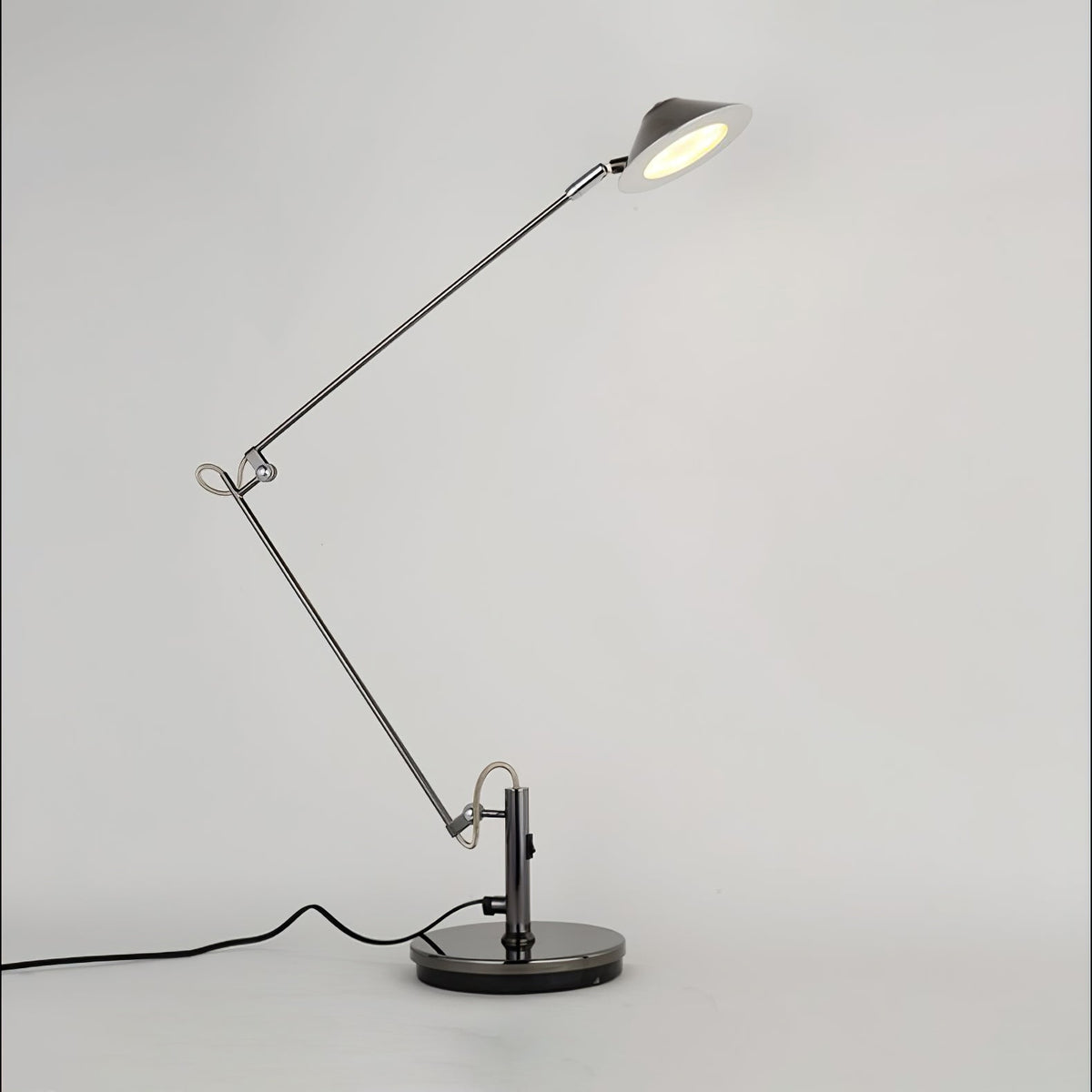 A Clérachic black arm lamp on a round base with the shade raised and lit, shown on a pale studio backdrop.