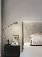 A Clérachic black arm lamp lights a black bedside table with two drawers, next to a framed print and a grey headboard.