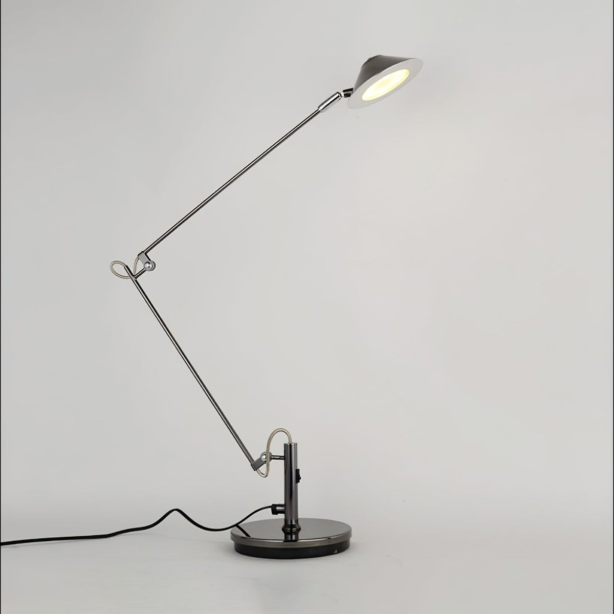 A Clérachic black arm lamp with a tilted, lit shade and a looped brass-toned joint, on a pale backdrop with a black cord.