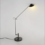 A Clérachic black arm lamp with a lit cone shade held high, a warm glow on the wall behind and a black cord on the floor.