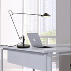 A Clérachic black arm lamp on a white desk, its lit shade reaching over a laptop, with a city view through the window.