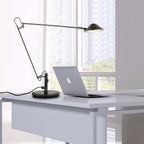 A Clérachic black arm lamp beside a laptop on a white desk, its arm held out and shade lit, with a view of city blocks.