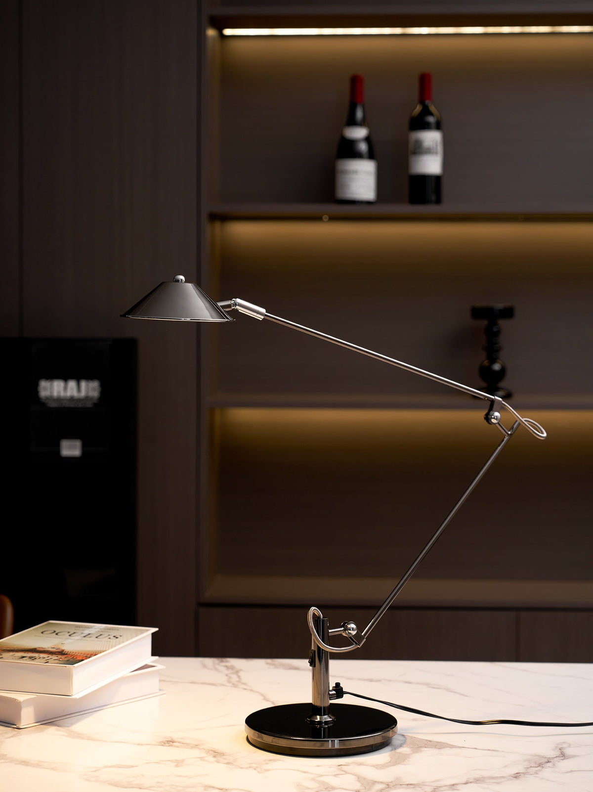 A Clérachic black arm lamp stands on a marble worktop, its shade held out in front of a dark shelving unit with wine bottles.