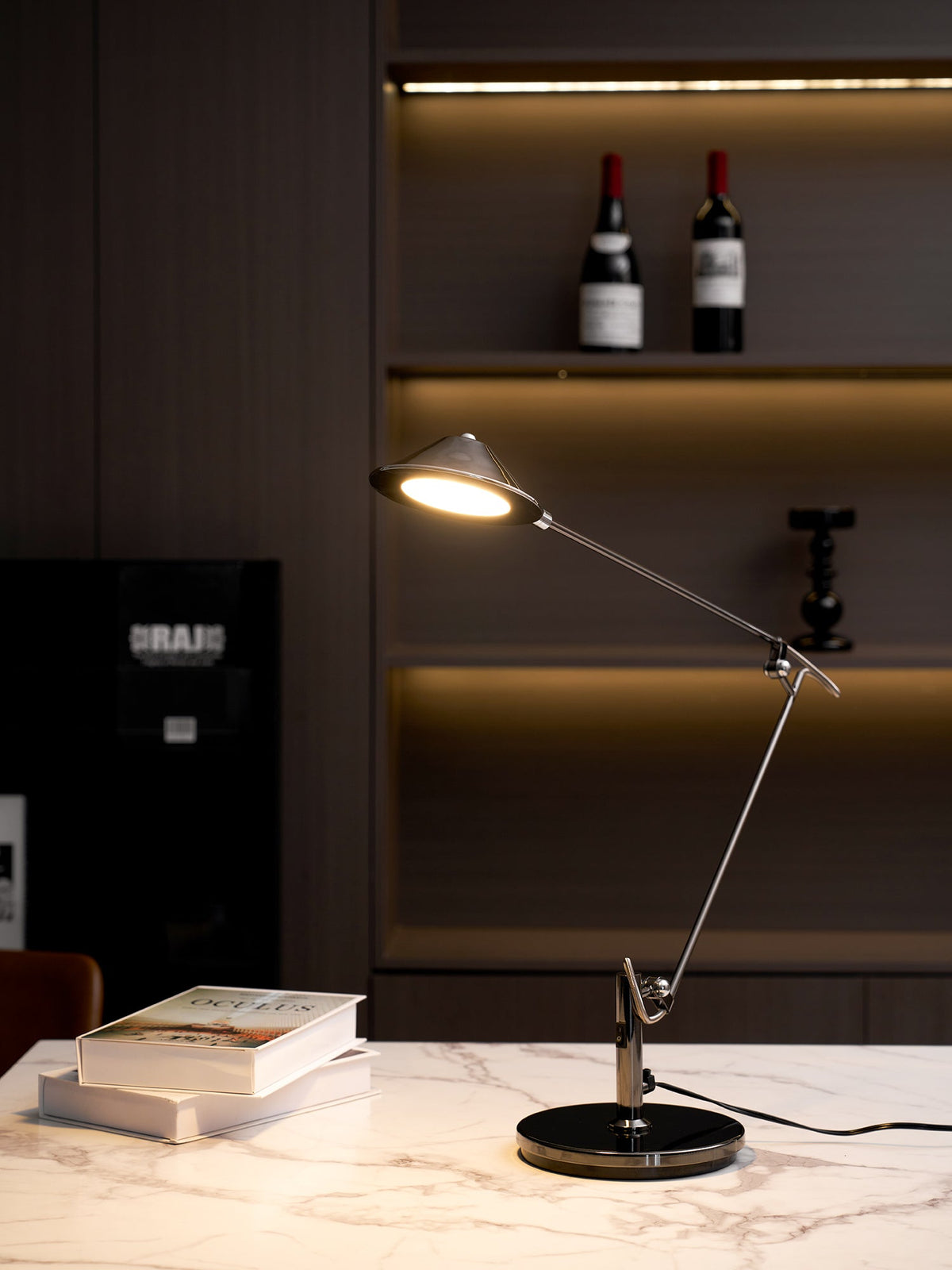 A Clérachic black arm lamp on a marble counter, lighting a stack of books, with warm shelf lighting and wine bottles behind.