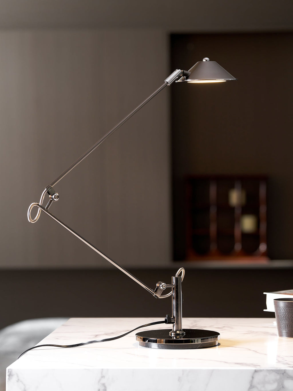 A Clérachic black arm lamp on a marble surface, its shade held high and lit, in front of a dark panelled wall and a clock.