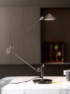 A Clérachic black arm lamp on a marble surface, its shade held high and lit, in front of a dark panelled wall and a clock.