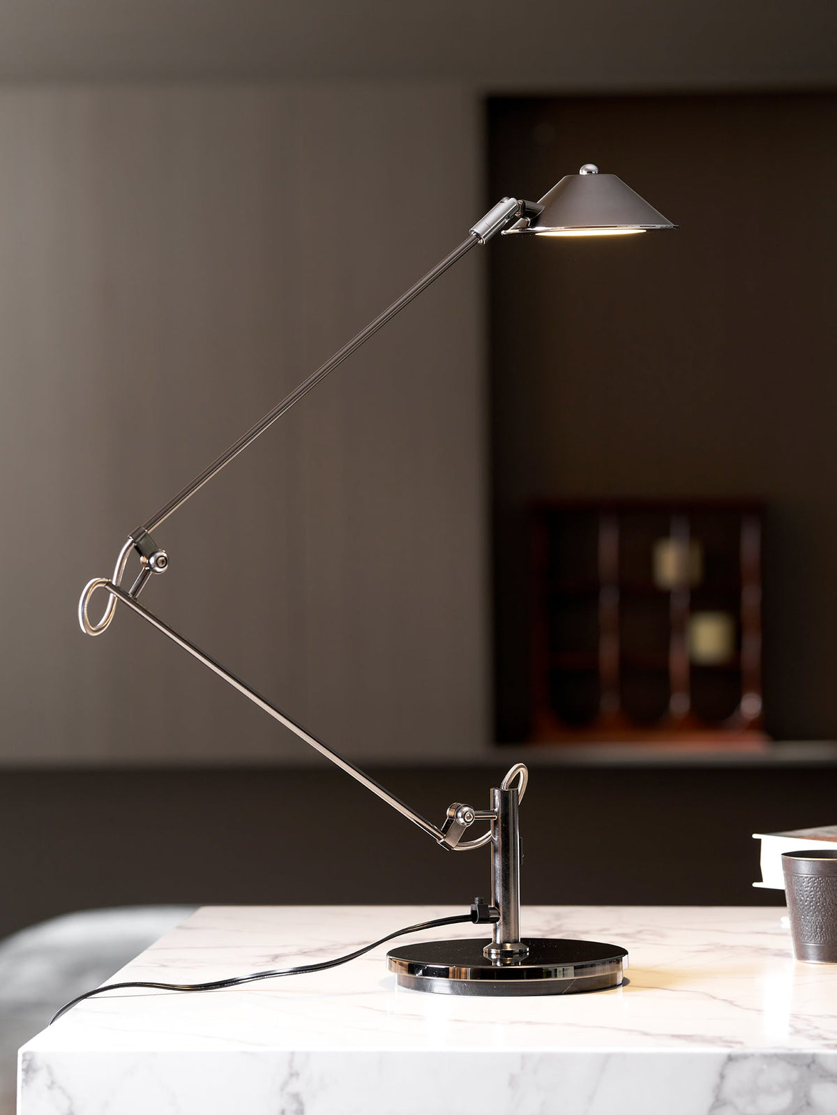 A Clérachic black arm lamp on a marble surface, its shade held high and lit, in front of a dark panelled wall and a clock.