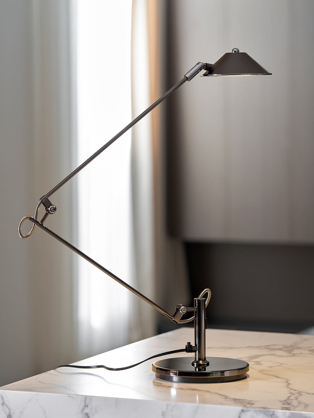 A Clérachic black arm lamp on a marble counter beside a window with sheer curtains, its arms folded into a wide angle.