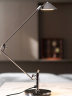 A Clérachic black arm lamp with a lit cone shade stands on a marble surface, with a bed and a wooden cabinet out of focus behind.