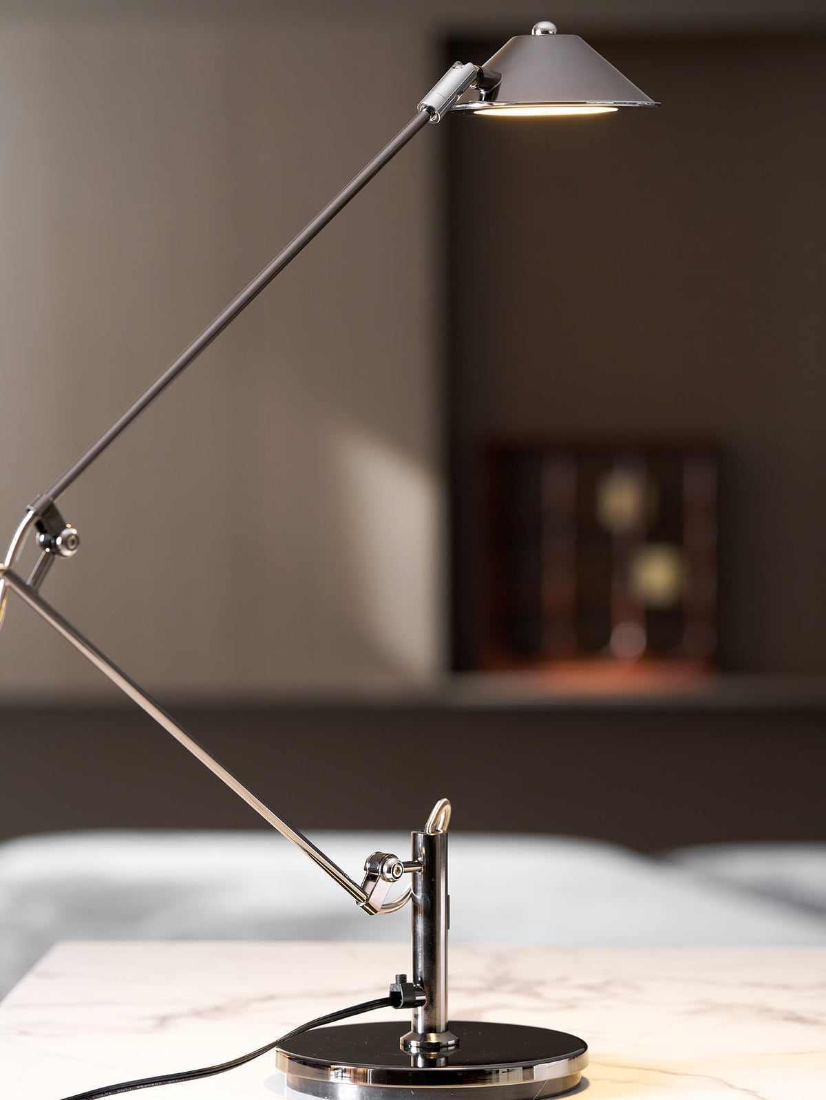 A Clérachic black arm lamp with a lit cone shade stands on a marble surface, with a bed and a wooden cabinet out of focus behind.