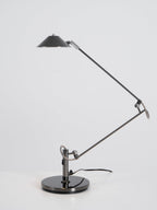 A Clérachic black arm lamp with its shade lowered over a table, shown unlit on a plain white-grey backdrop.