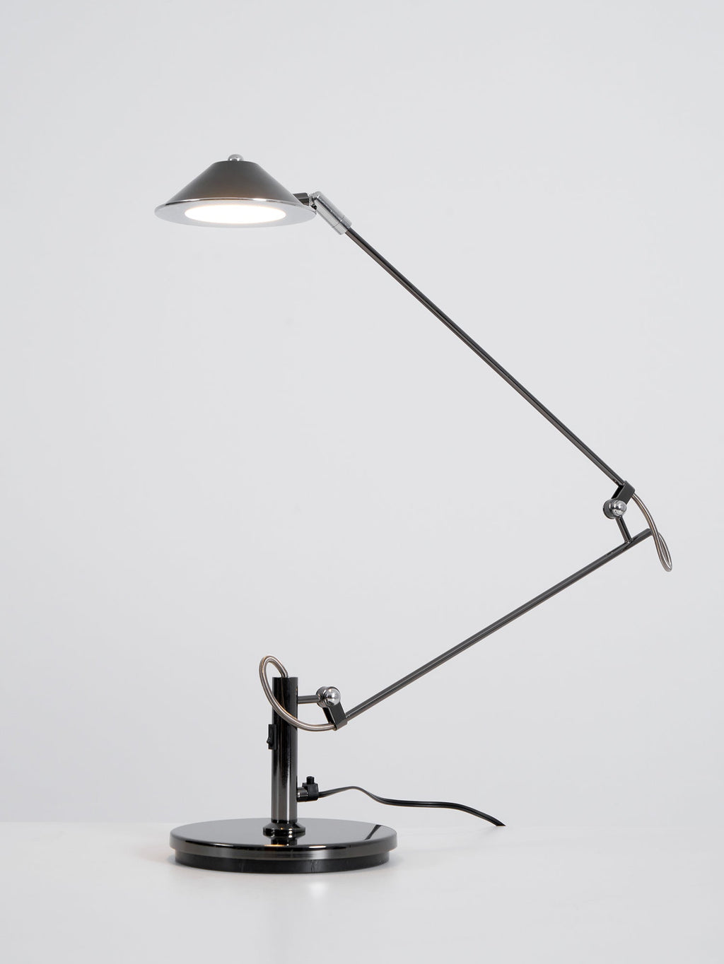 A Clérachic black arm lamp with its shade raised and the LED switched on, on a plain white-grey backdrop.