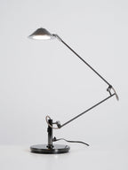 A Clérachic black arm lamp with its shade raised and the LED switched on, on a plain white-grey backdrop.