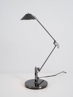 A Clérachic black arm lamp with a folded arm and a high, unlit cone shade, standing on a white surface against grey.