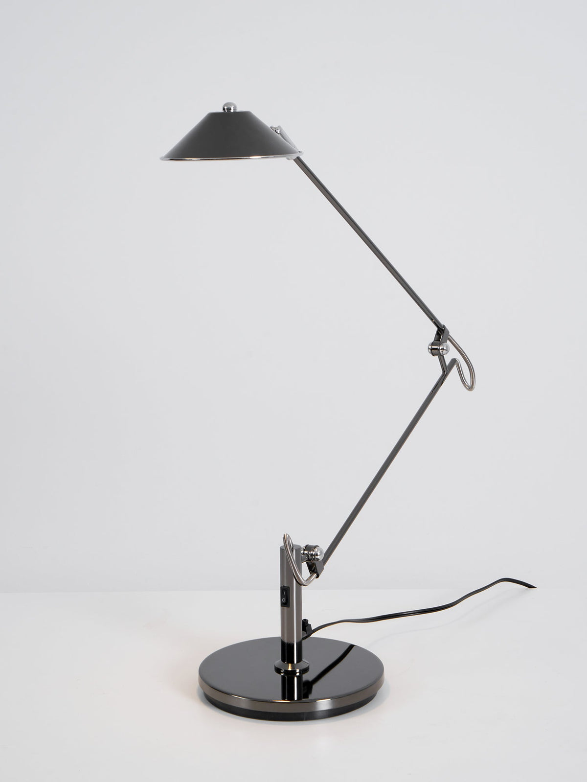 A Clérachic black arm lamp with a folded arm and a high, unlit cone shade, standing on a white surface against grey.