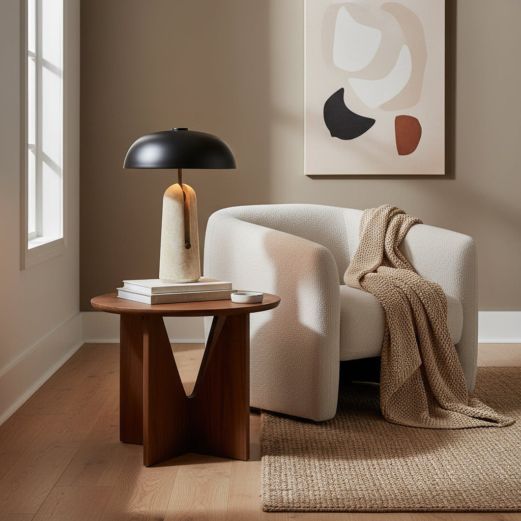 On a round walnut side table beside a cream armchair, this Clérachic table lamp sits under a window with an abstract print behind.