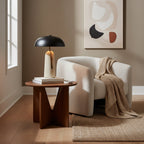 On a round walnut side table beside a cream armchair, this Clérachic table lamp sits under a window with an abstract print behind.