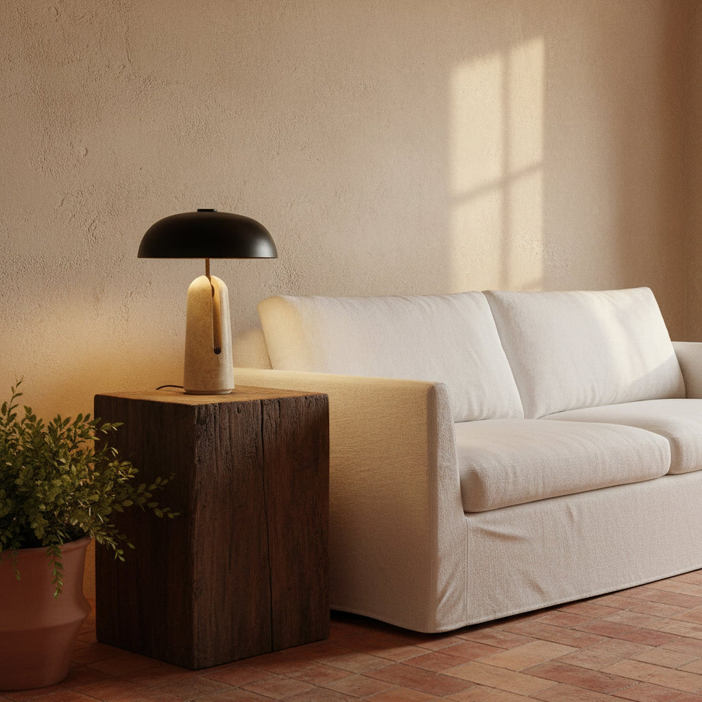 Warm light from this Clérachic table lamp on a dark timber block beside a white sofa, with a potted plant at the side.