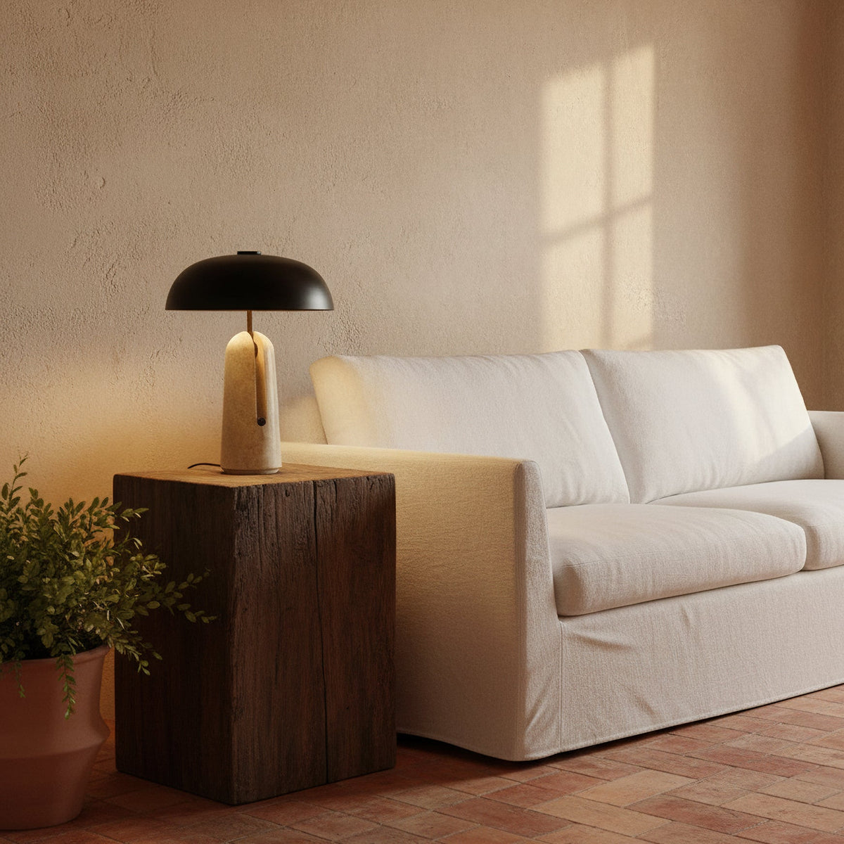Warm light from this Clérachic table lamp on a dark timber block beside a white sofa, with a potted plant at the side.