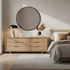 A Clérachic table lamp on a light oak chest of drawers under a round black-framed mirror, with a bed at the right.